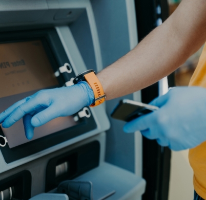 Cropped shot of man wears protective rubber gloves, uses ATM machie to withdraw money, prevents from corona virus or covid-19, holds mobile phone, presses button. Virus pandemic. Health care concept
